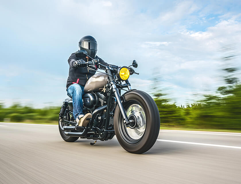 A person in a black helmet rides a classic-style motorcycle with a yellow headlight on an open road, enjoying the thrill of speed. The clear blue sky and blurred greenery whiz by, reminding one of lifes fleeting moments. Dont forget your auto insurance for peace of mind on every journey.